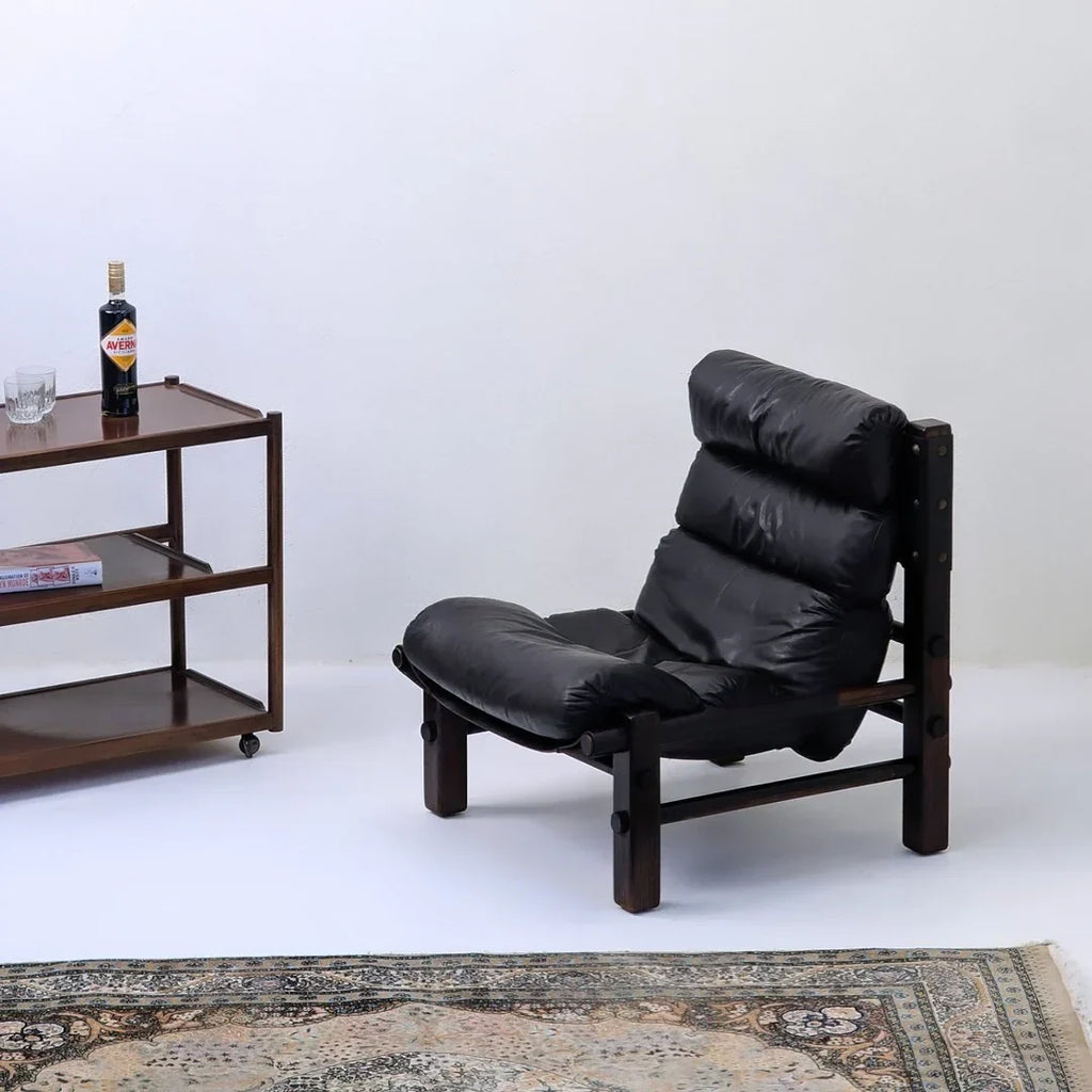 Vintage black leather lounge chair with wooden frame, bar cart, and patterned rug in minimalist room