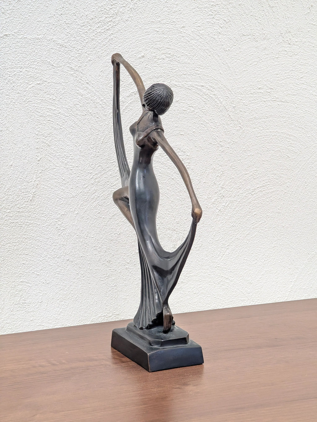 bronze sculpture of a dancing woman in a flowing dress on a wooden surface with textured white wall background