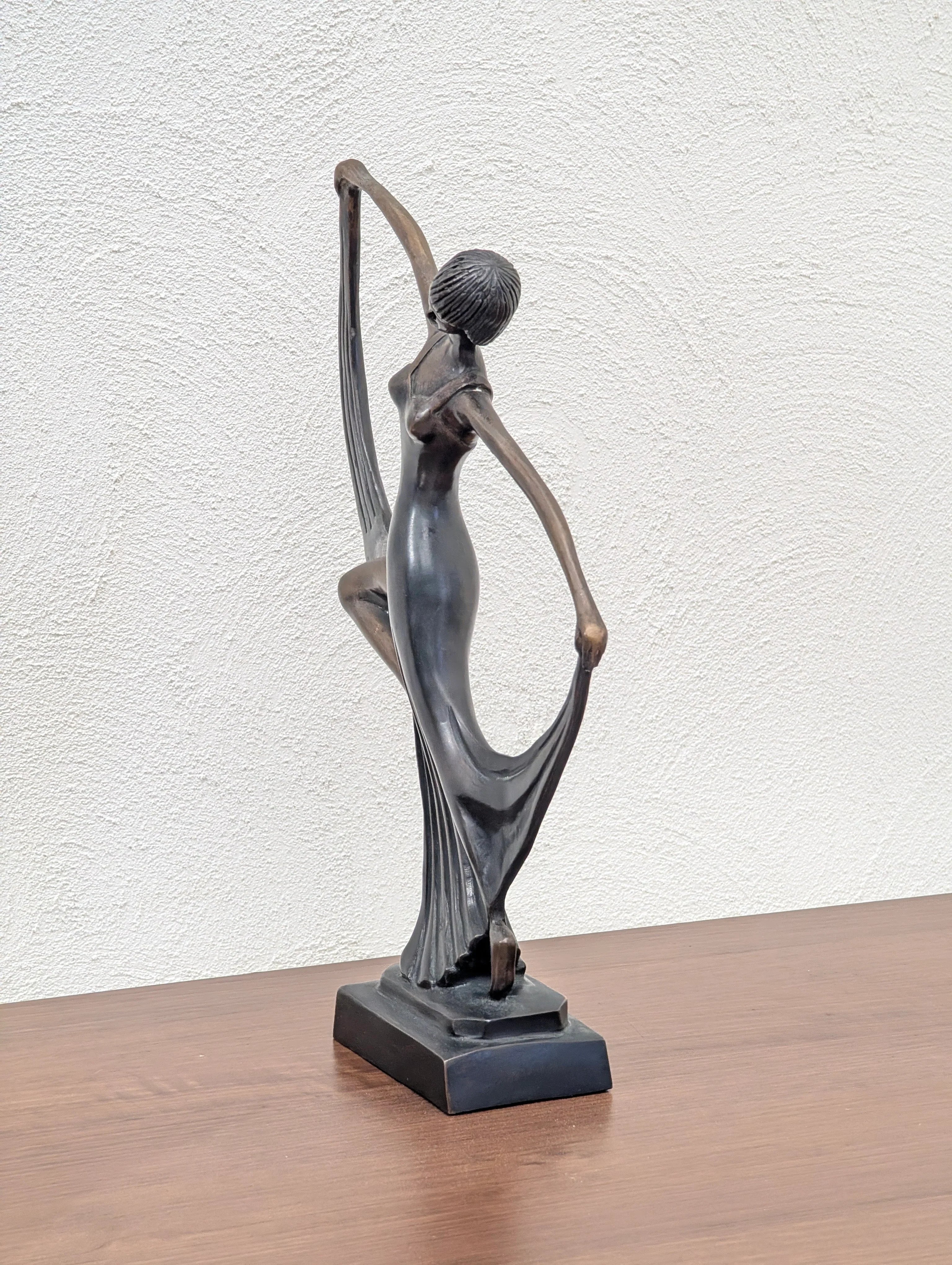 bronze sculpture of a dancing woman in a flowing dress on a wooden surface with textured white wall background