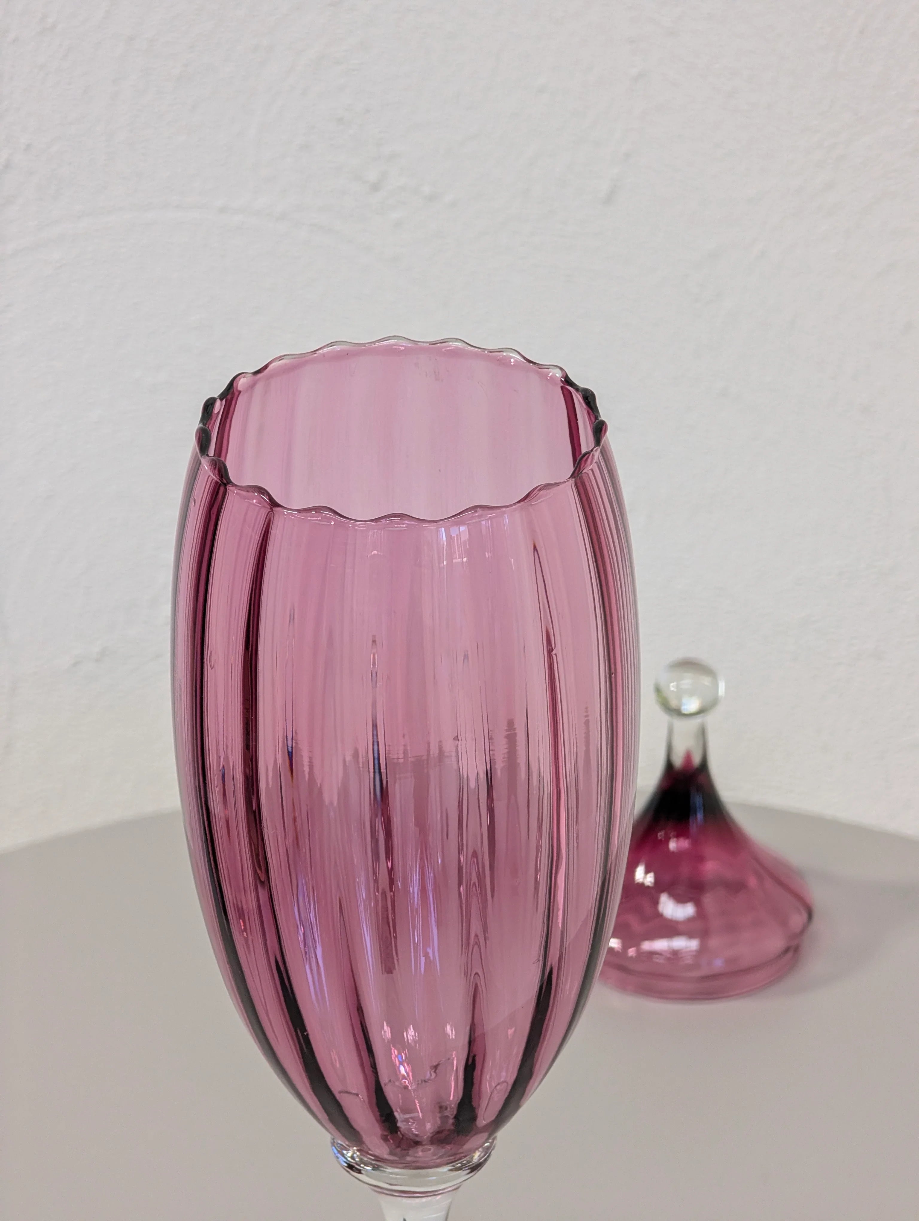 Close-up of tall pink ribbed glass vase with scalloped rim and matching lid on white surface