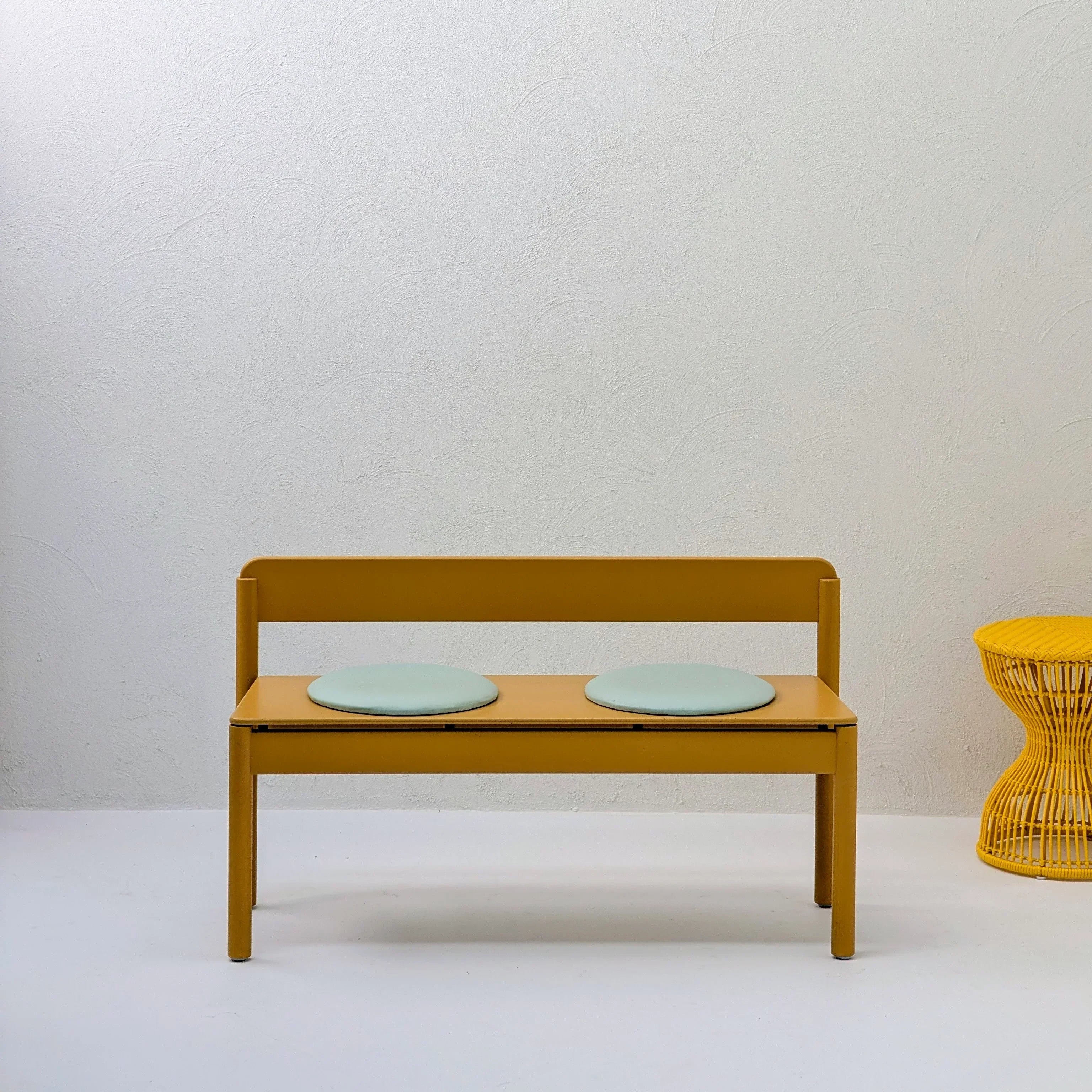 Minimalist mustard yellow bench with two round cushions, next to a yellow woven stool, white wall