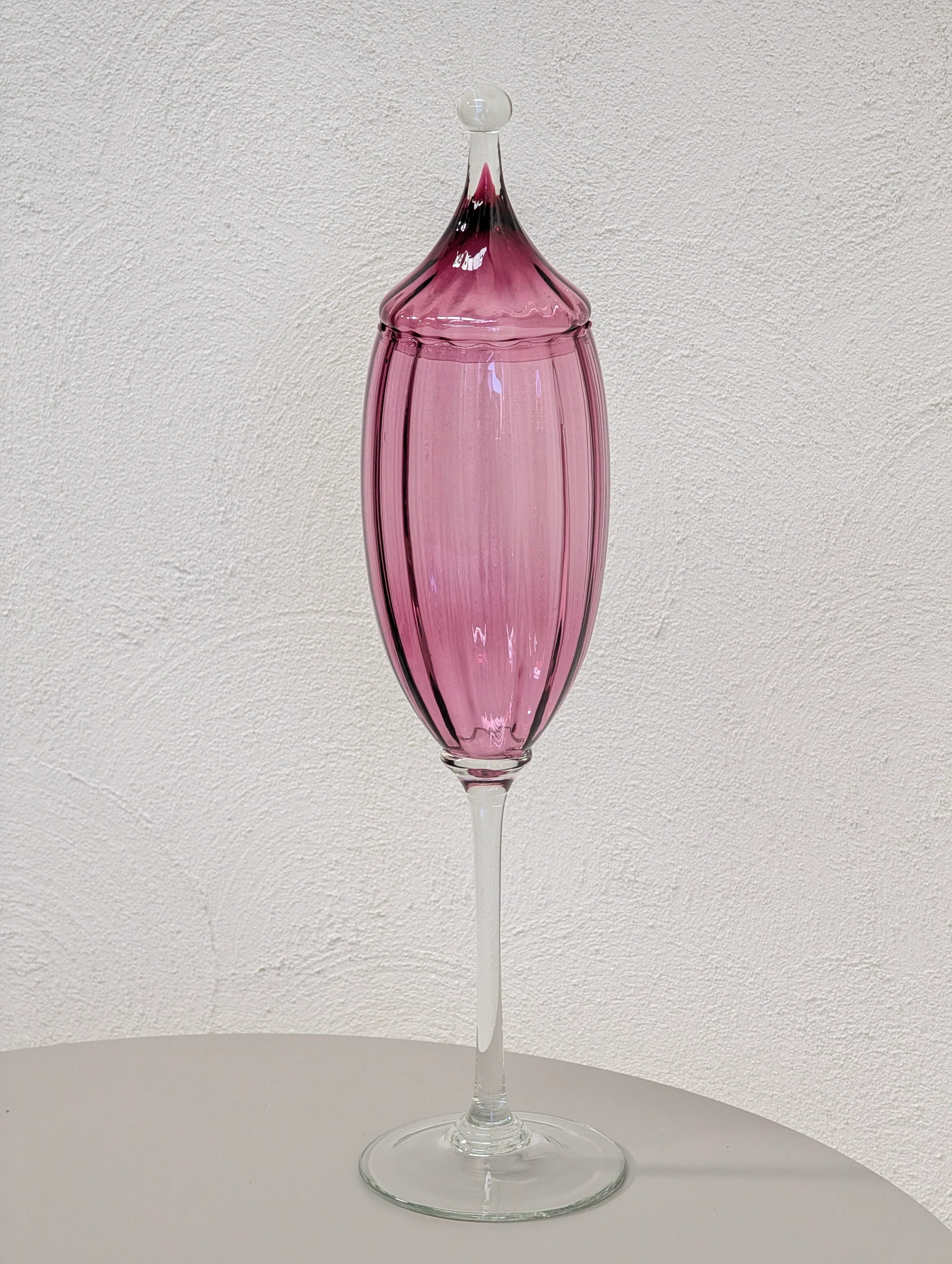Tall pink glass decanter with a pointed lid on a round white table against a white textured wall