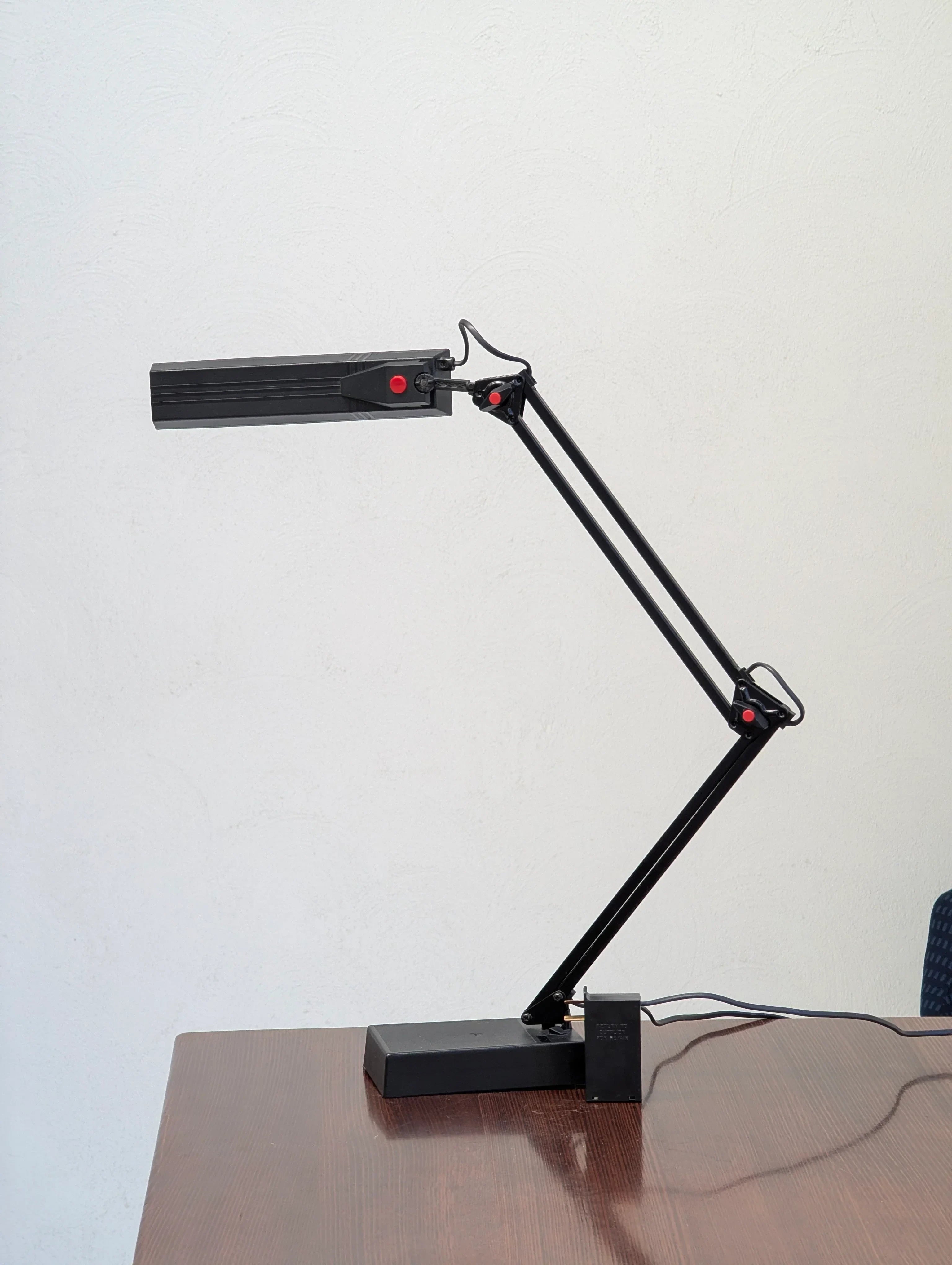 Black adjustable desk lamp on a wooden table against a white wall, plugged in with a power adapter.