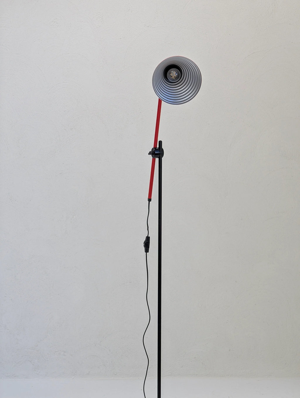Modern floor lamp with red and black stand against a plain textured white wall