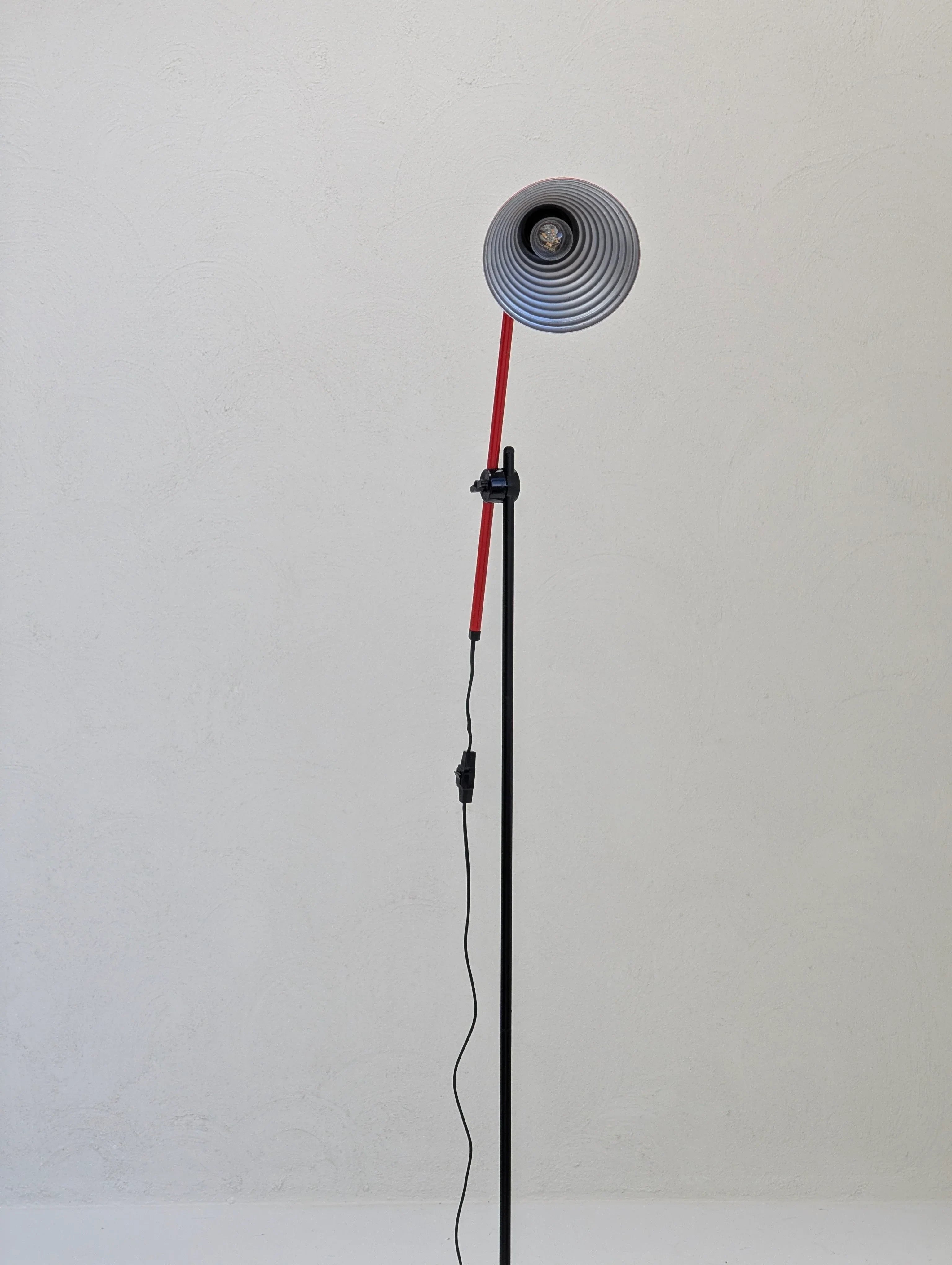 Modern floor lamp with red and black stand against a plain textured white wall