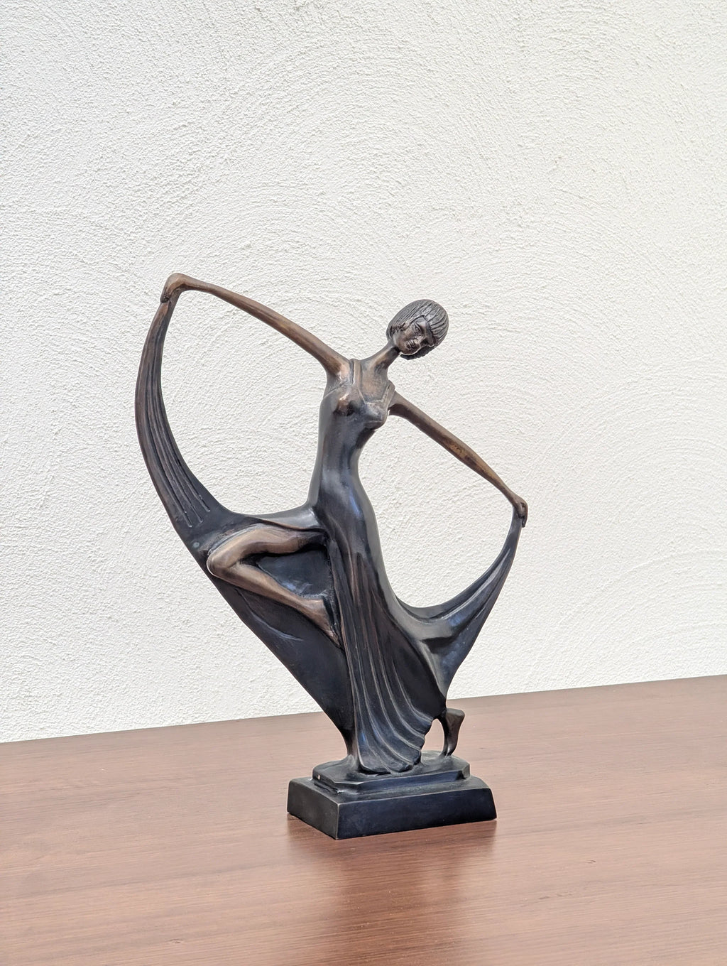 Bronze sculpture of a dancing woman in a flowing dress on a wooden surface against a white textured wall