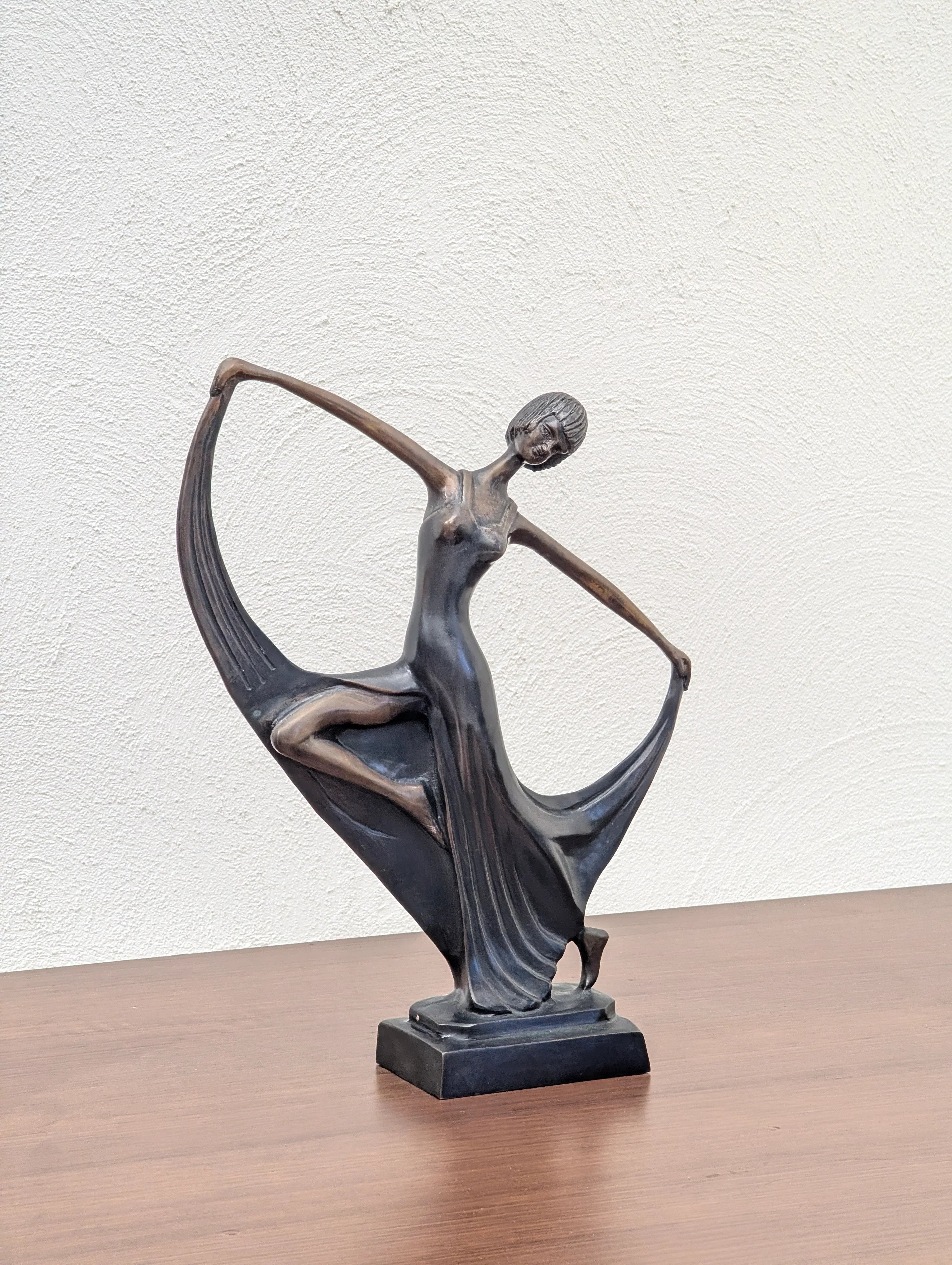 Bronze sculpture of a dancing woman in a flowing dress on a wooden surface against a white textured wall