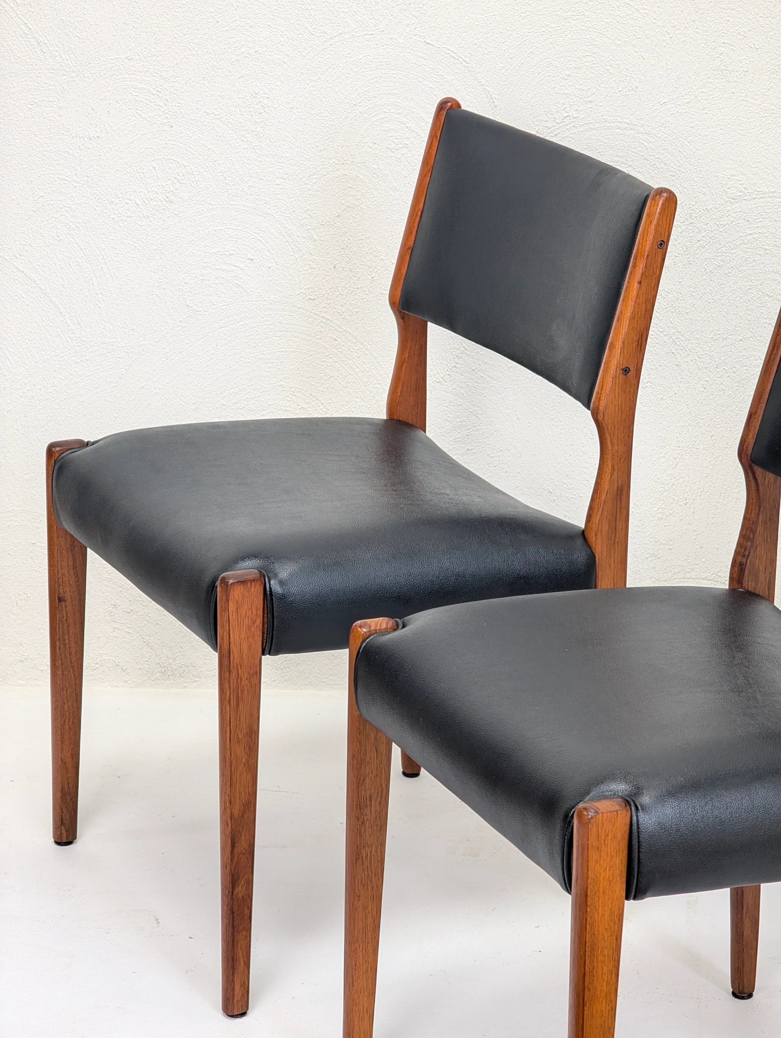 Two mid-century modern dining chairs with black leather seats and teak wood frames against white wall