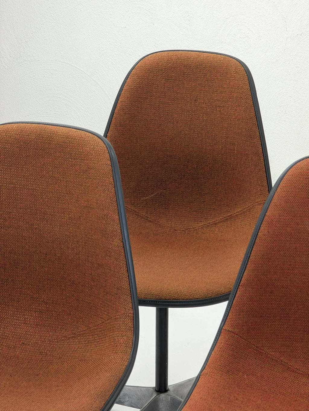 Three mid-century modern orange upholstered swivel chairs with black trim against a white wall