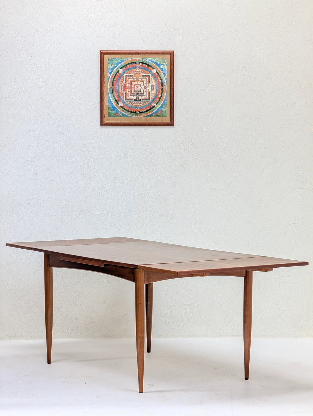 Wooden mid-century modern dining table with tapered legs in minimalist white room beneath colorful framed artwork