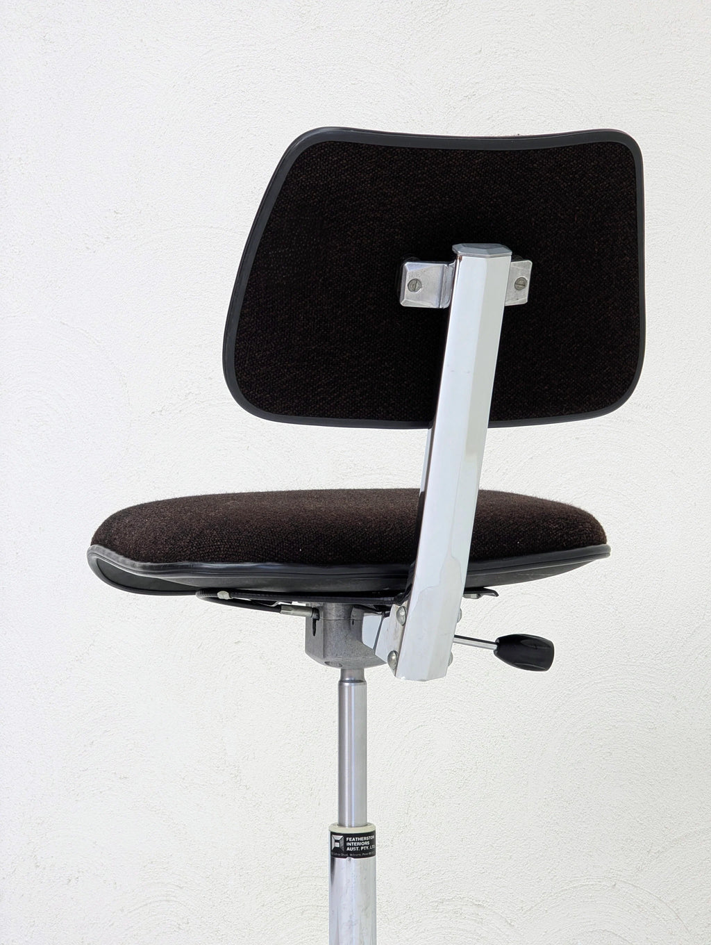 Black cushioned office chair with chrome metal frame and adjustable height lever against white wall