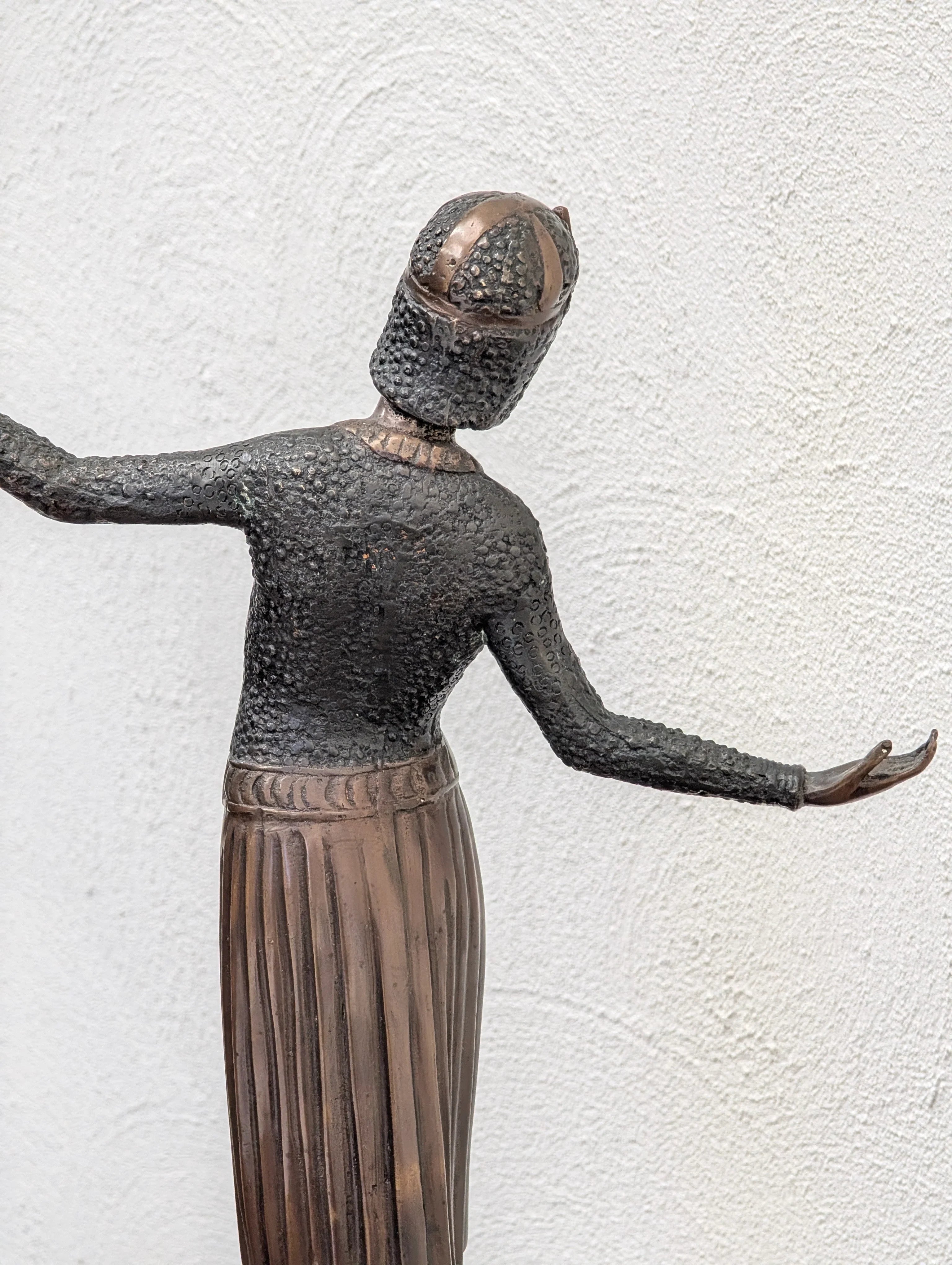 Bronze sculpture of a person with textured upper body and pleated skirt against a white wall
