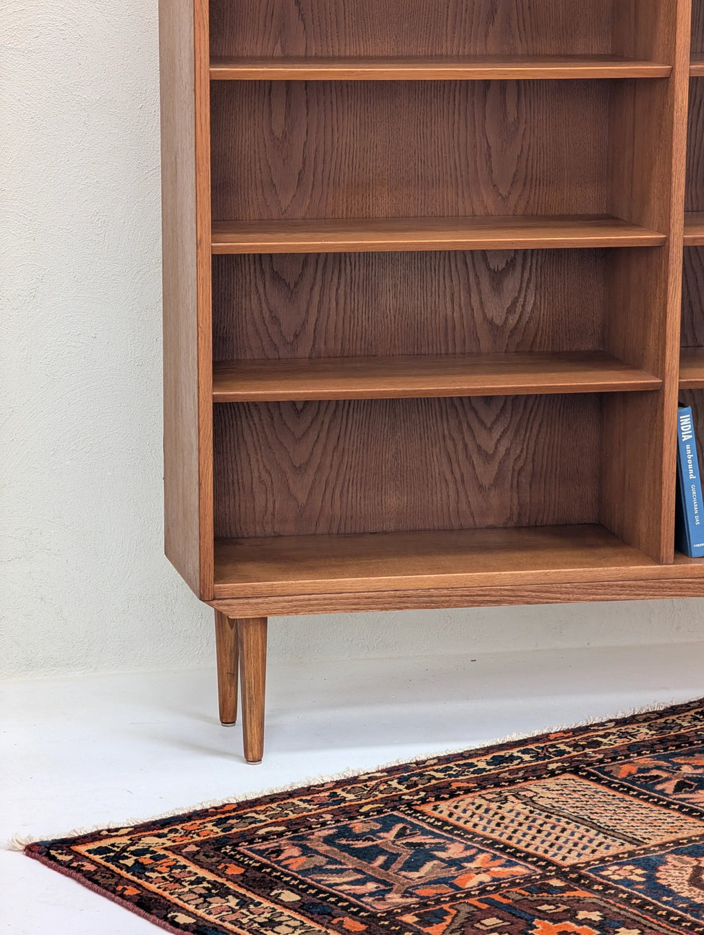 Mid-century wooden bookshelf with tapered legs on patterned vintage rug and white wall background