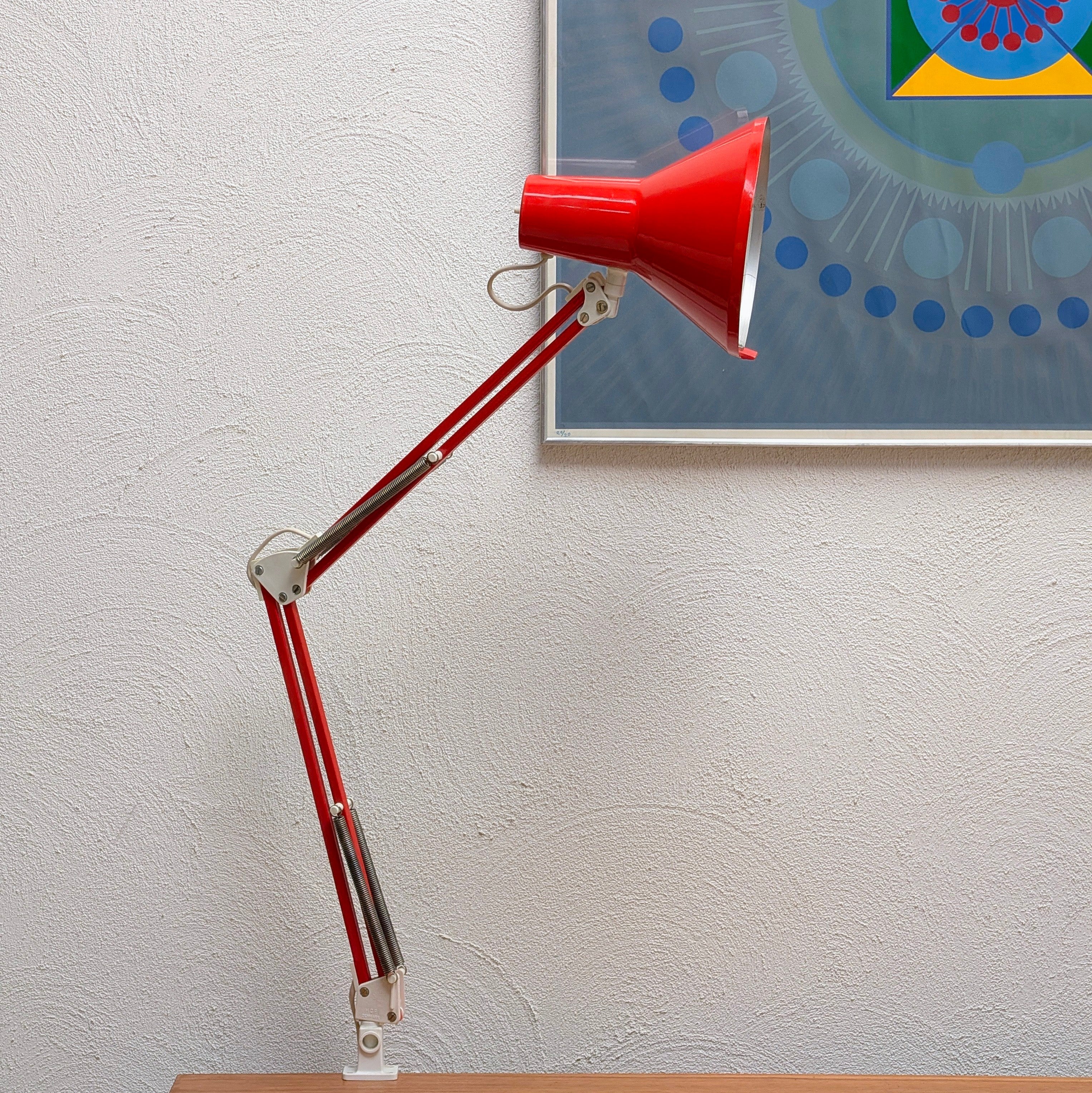 A red adjustable desk lamp with a cone-shaped shade and articulated arm, shown on a tabletop against a textured white wall with a framed abstract print partially visible behind it.