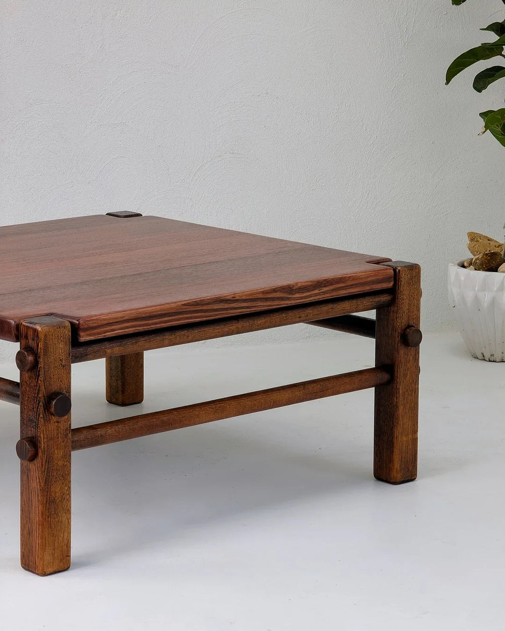 Rustic wooden coffee table with thick legs and exposed joinery against white wall with potted plant