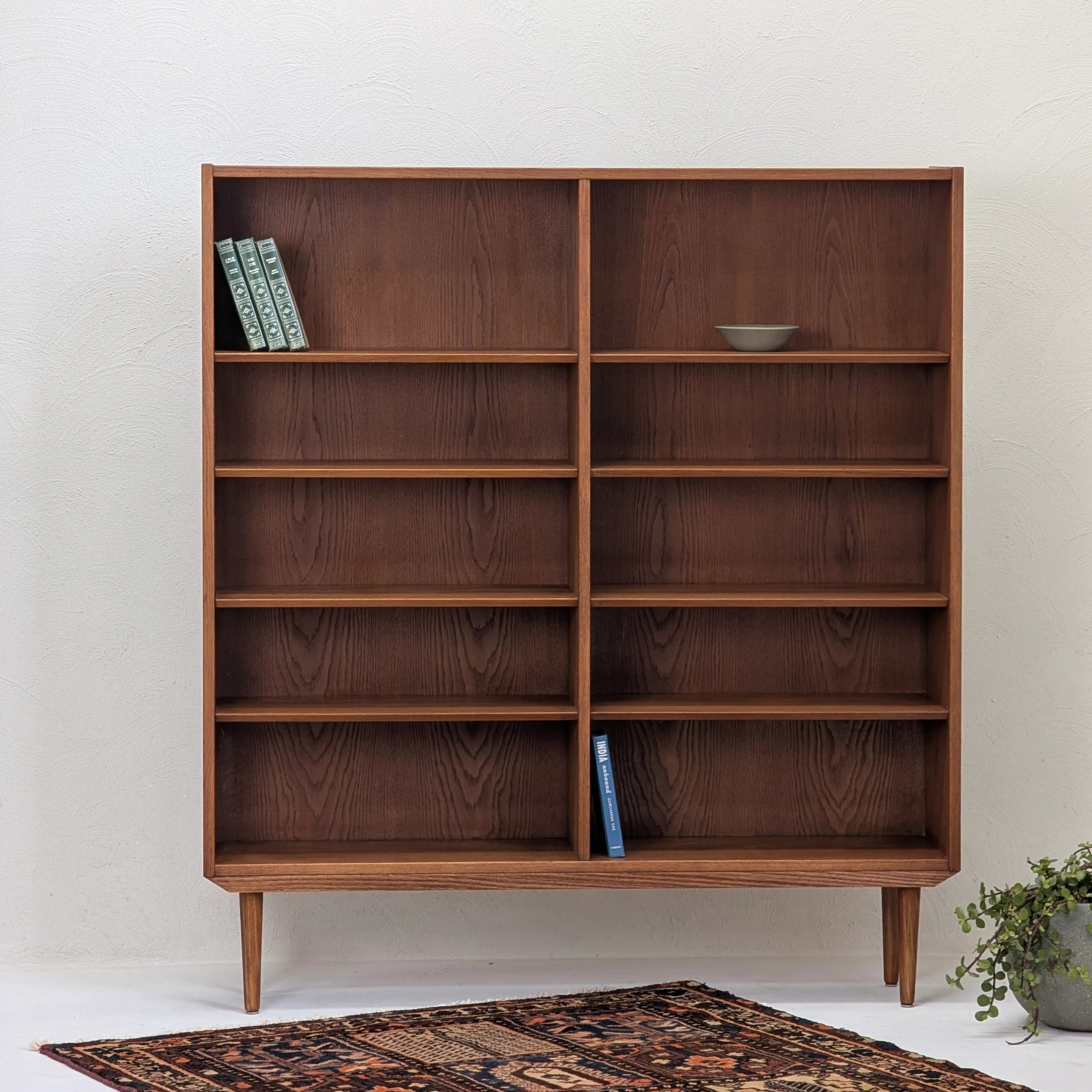 Mid-century modern wooden bookshelf with tapered legs, sparsely decorated with books and ceramic bowl, vintage rug and plant nearby
