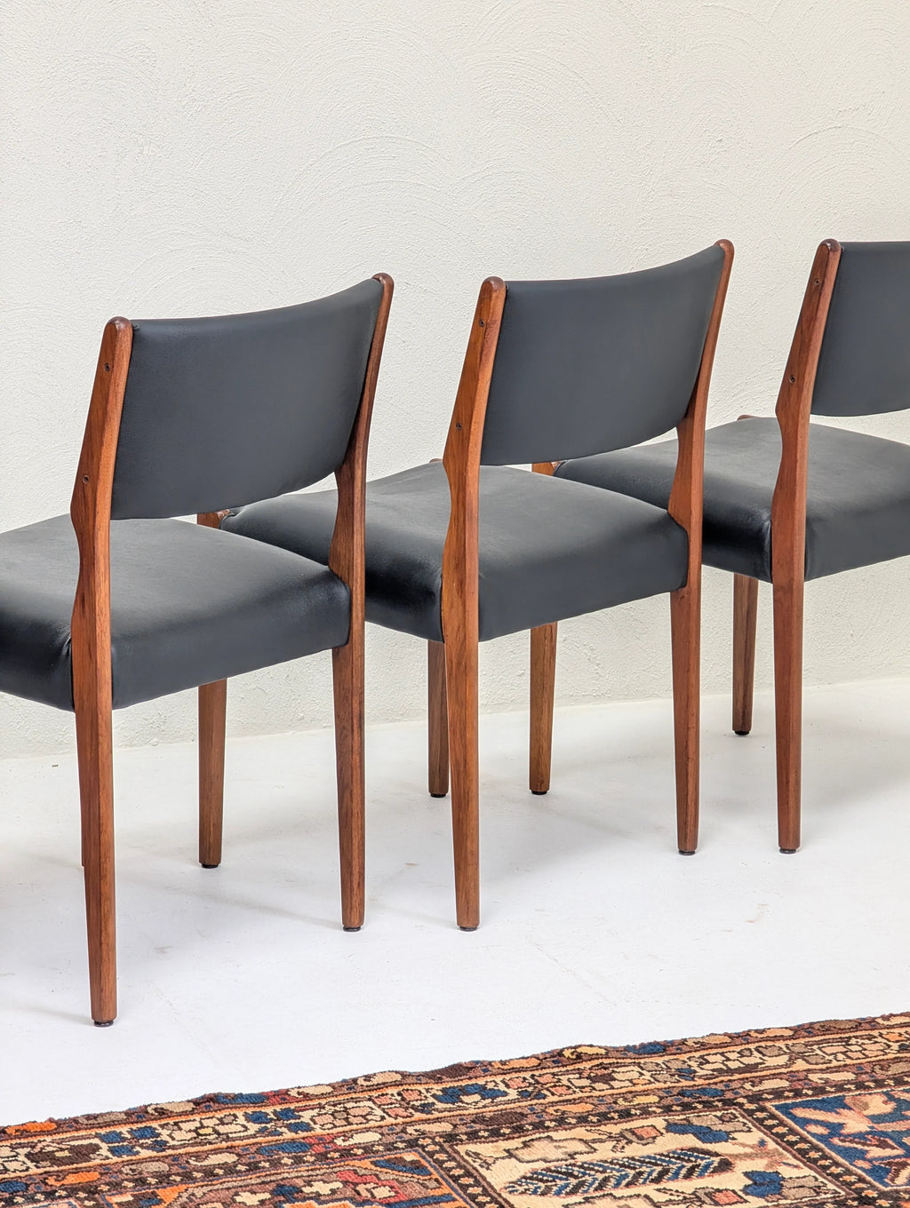 Three mid-century modern dining chairs with dark leather seats and wooden frames on white floor with patterned rug
