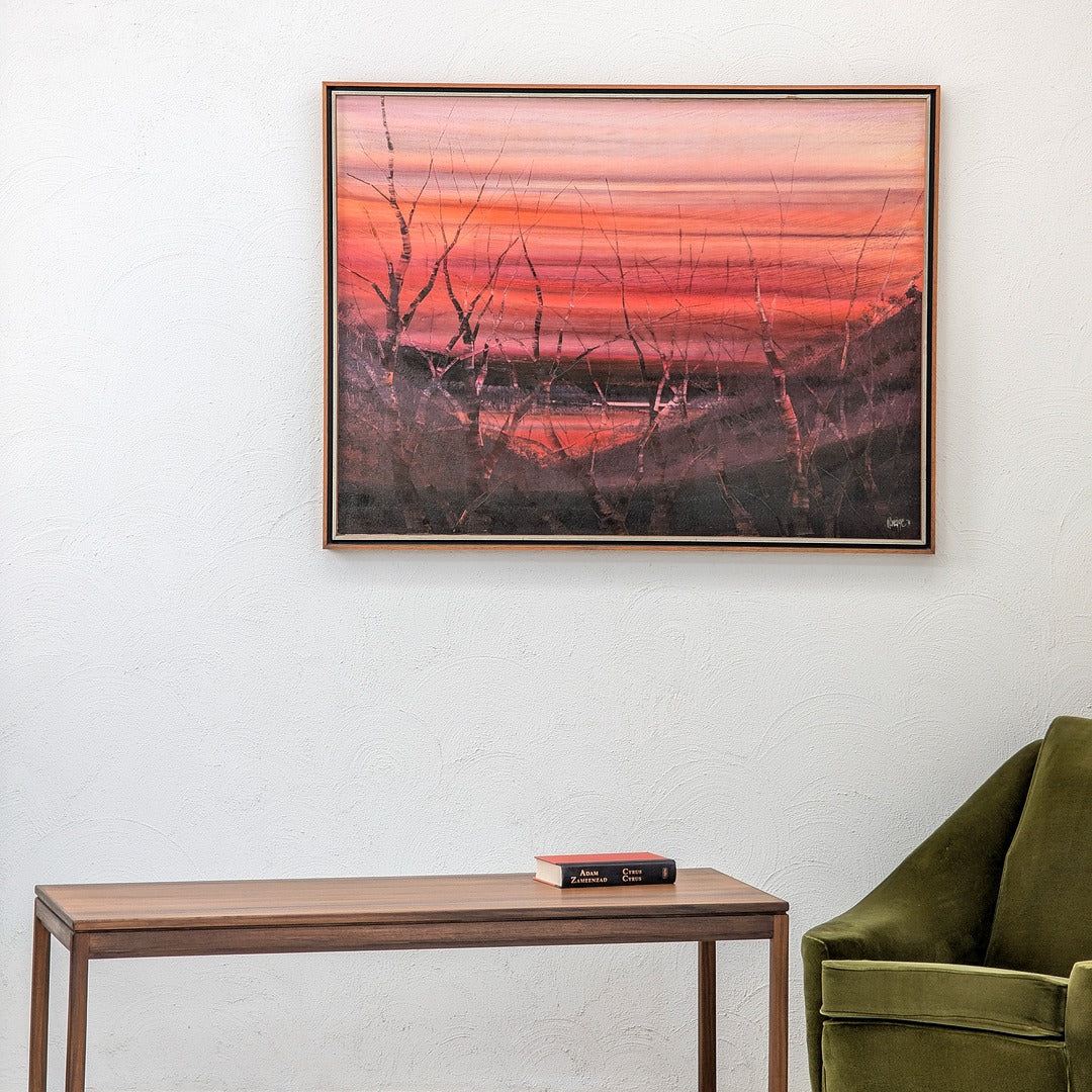 Framed sunset landscape painting on white wall above wooden table and green velvet chair
