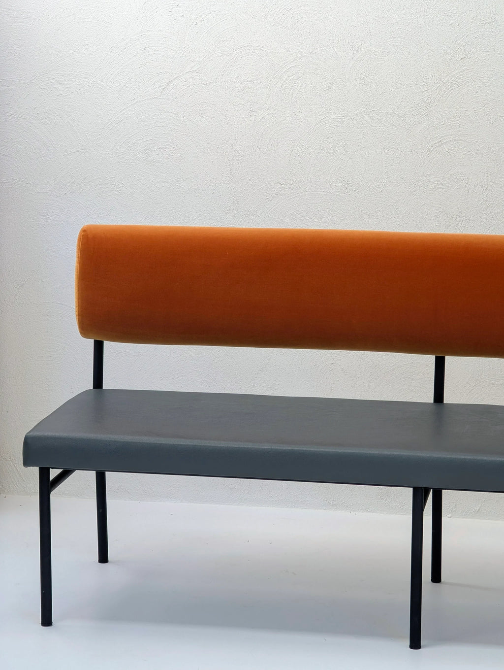 Modern bench with orange backrest, gray seat, black metal legs against textured white wall
