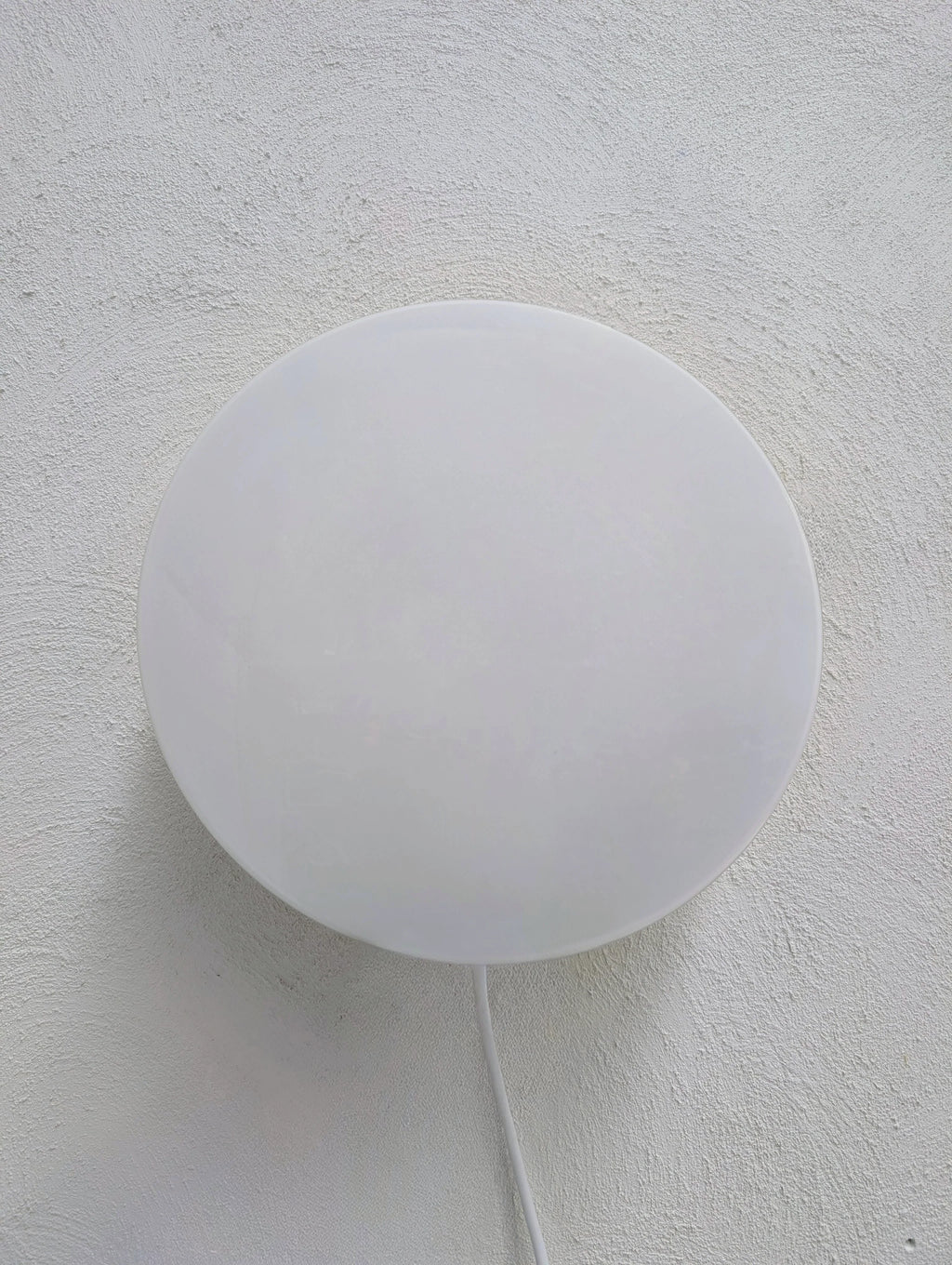 Minimalist white circular wall lamp mounted on textured white wall with cable hanging down