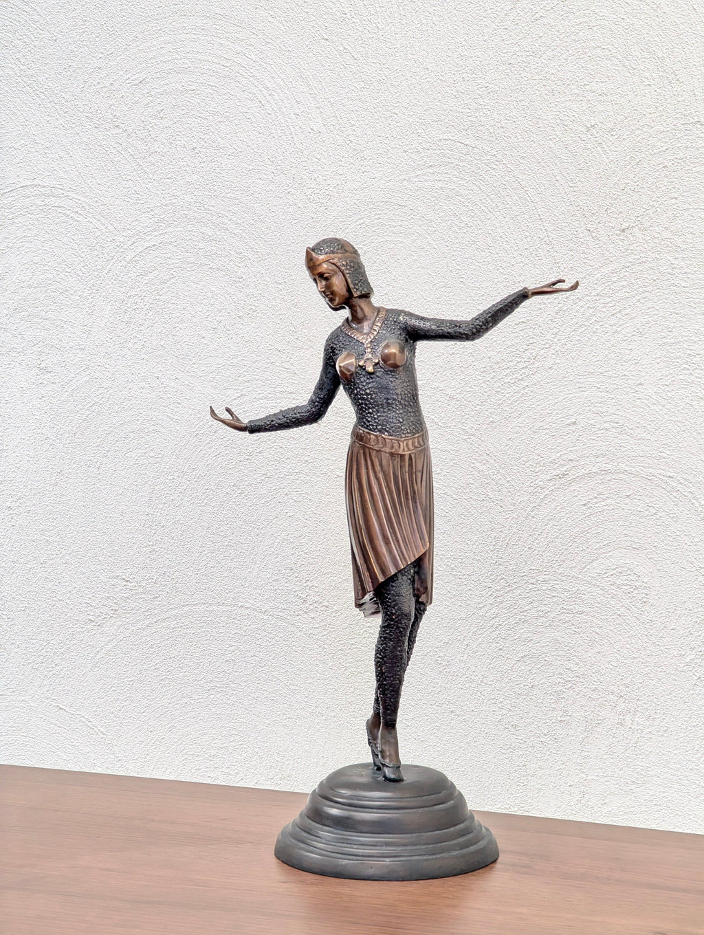 Bronze statue of a woman in a dance pose wearing a textured top, skirt, and heeled shoes on a black circular base
