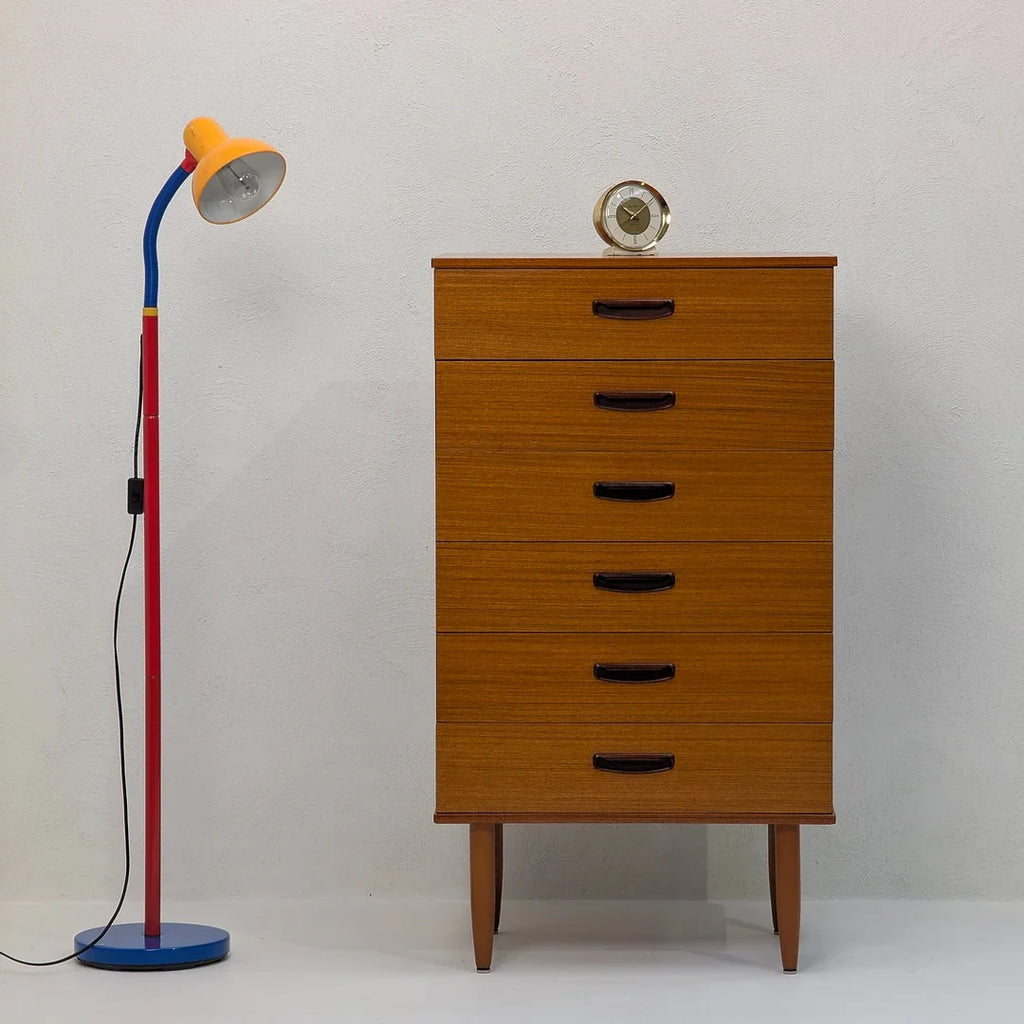 Mid-century wooden dresser with vintage clock beside colorful floor lamp against white wall