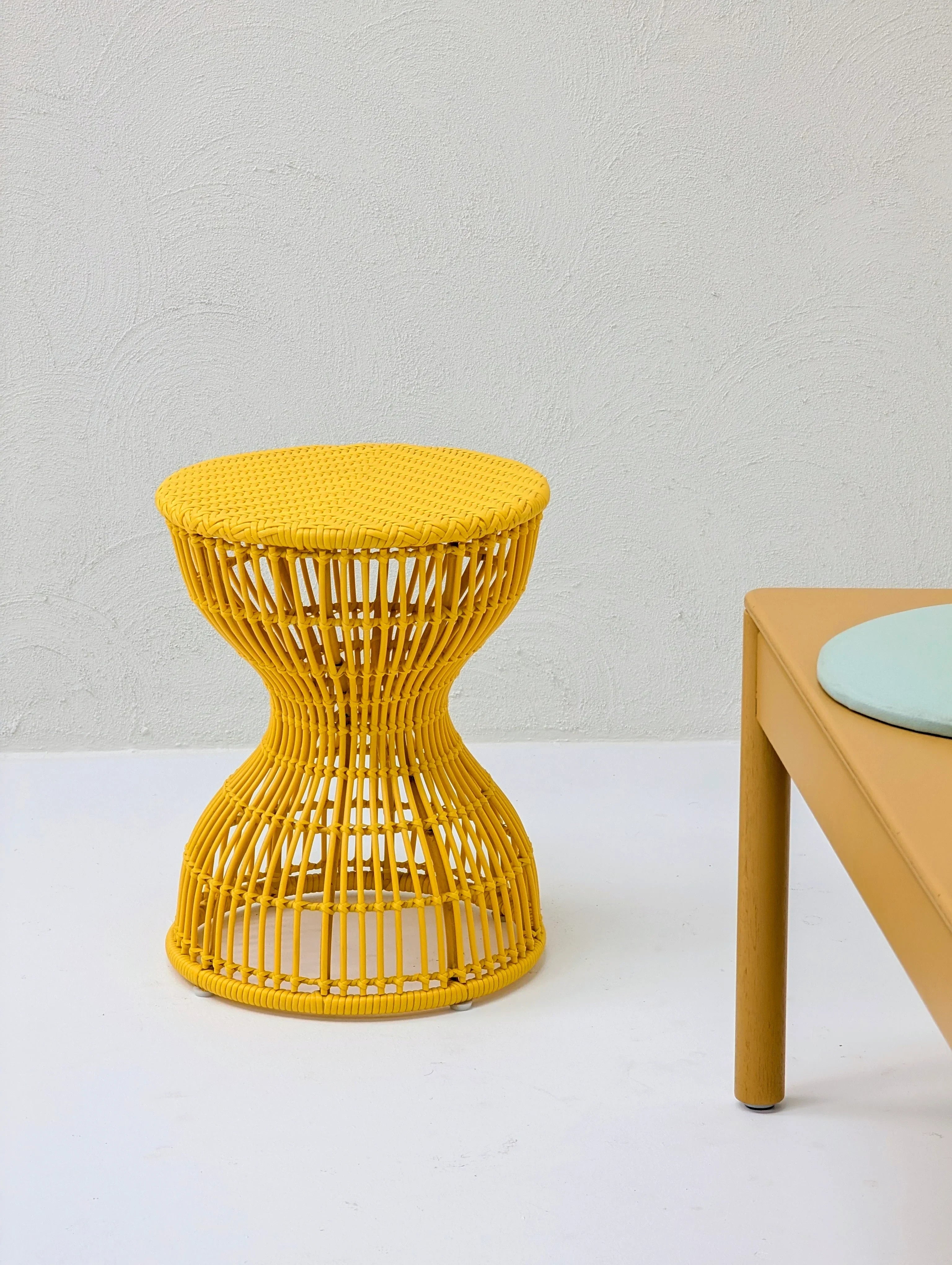 Yellow wicker stool beside a beige table with a round blue cushion on a white floor