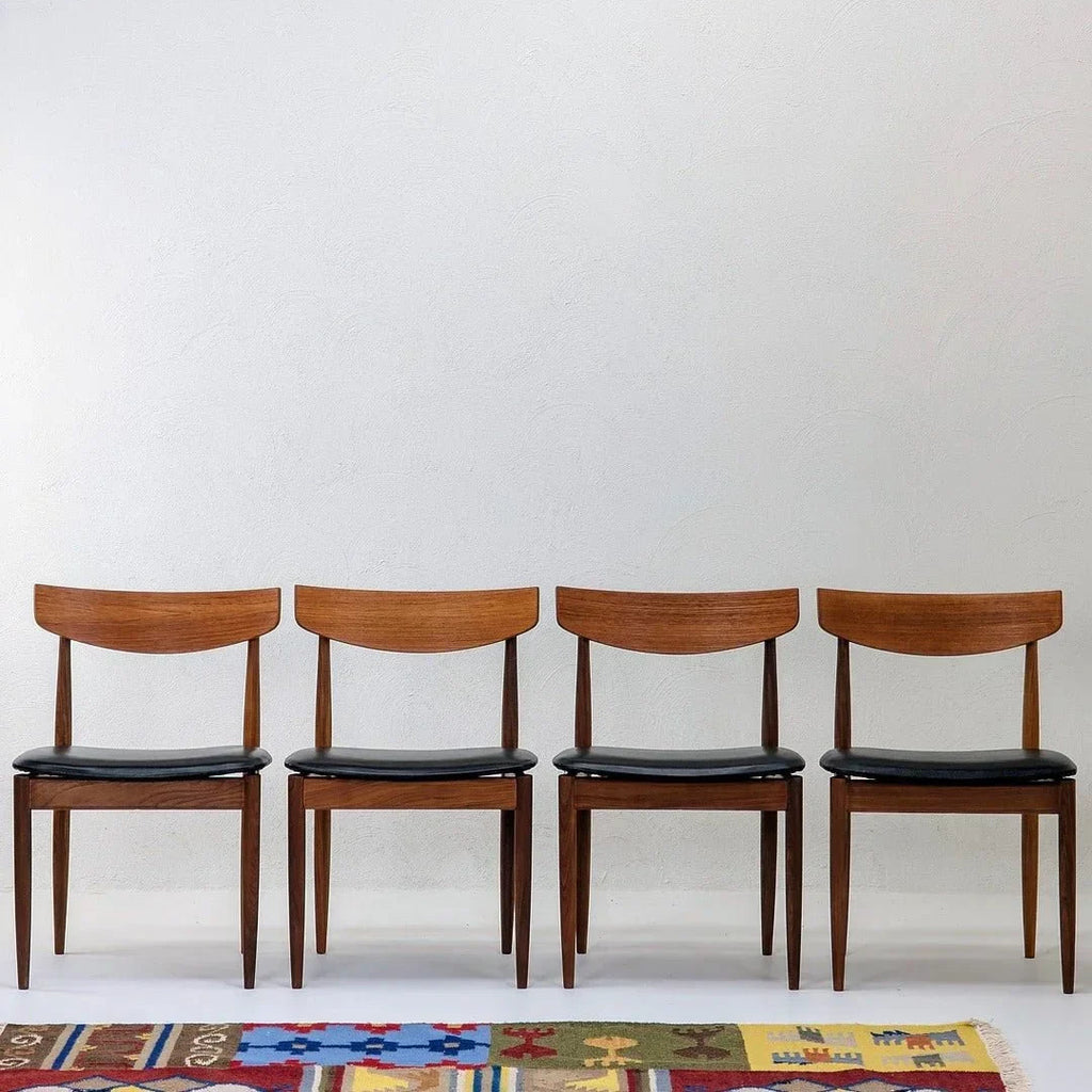 Set of four teak dining chairs with black seats, mid-century modern style, against white wall.