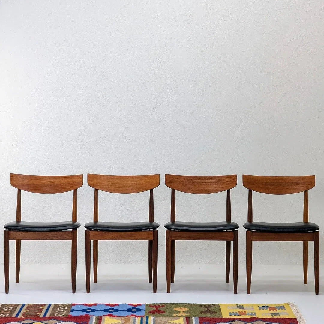 Set of four teak dining chairs with black seats, mid-century modern style, against white wall.
