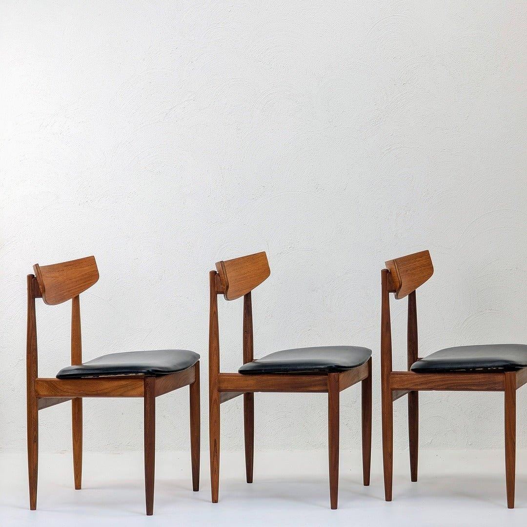 Set of three mid-century teak dining chairs with black leather seats against white wall