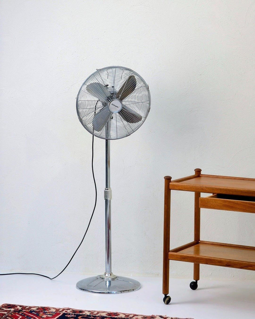 Vintage Dimplex stainless steel pedestal fan beside wooden cart on white floor