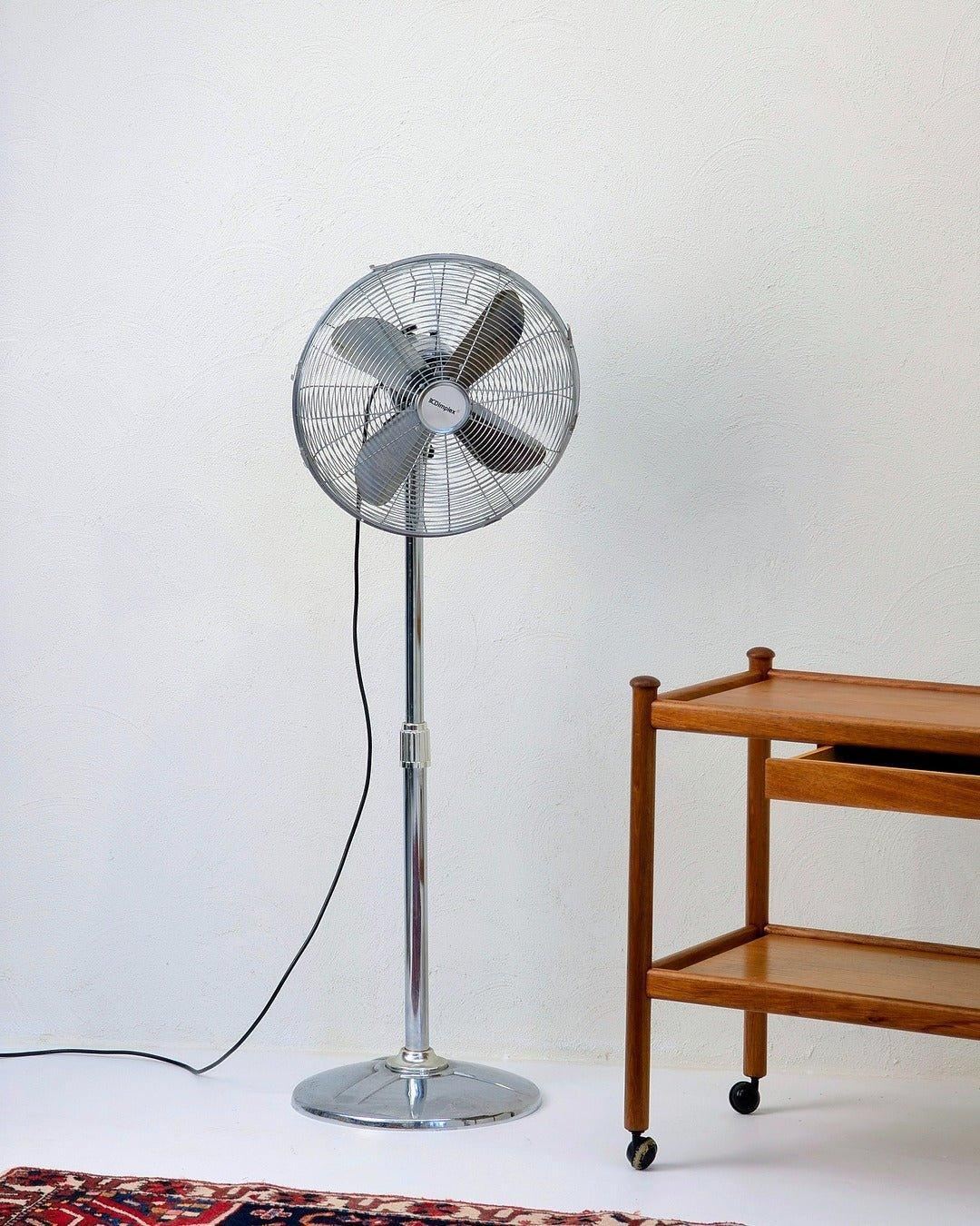 Vintage Dimplex stainless steel pedestal fan beside wooden cart on white floor