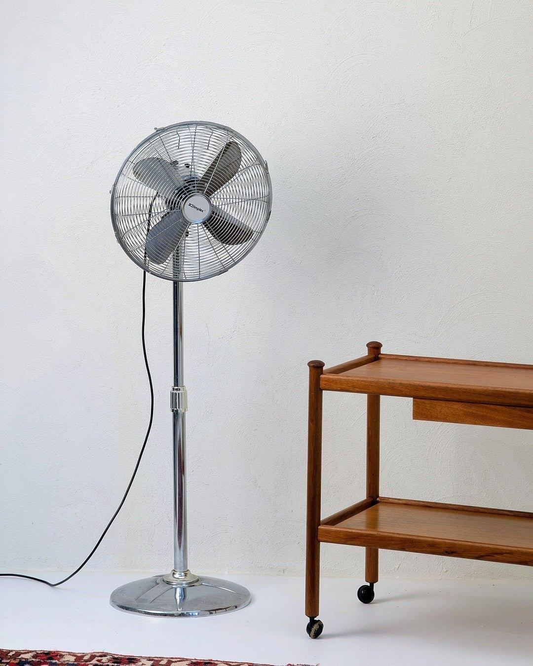 Vintage Dimplex stainless steel pedestal fan beside wooden cart on white background