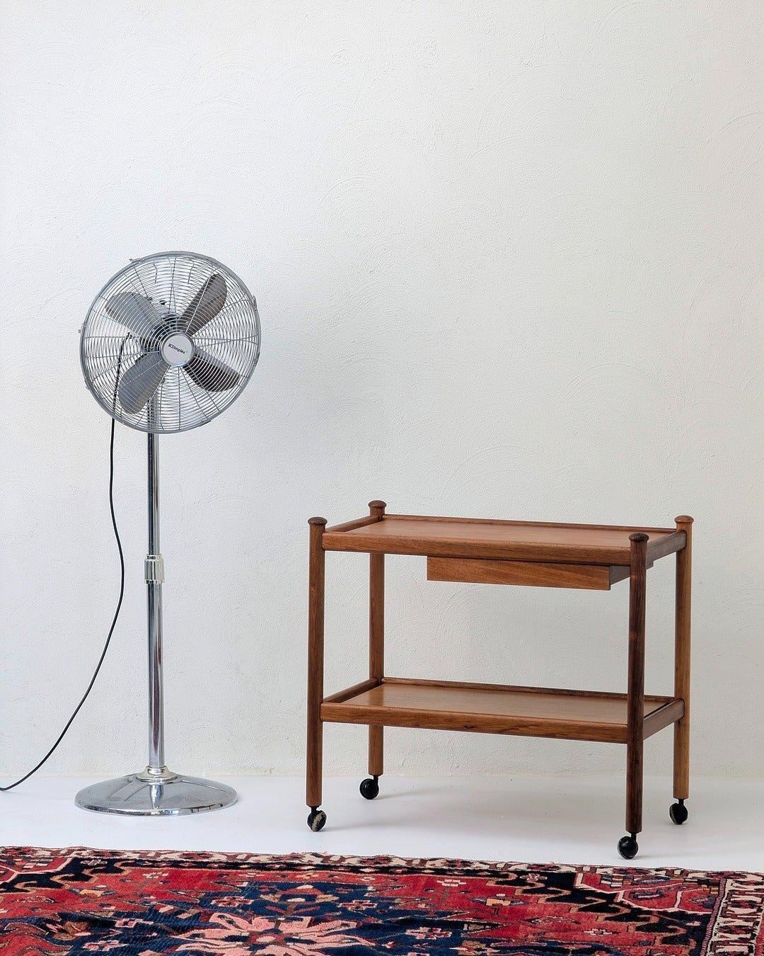 Vintage stainless steel pedestal fan next to wooden cart on wheels, patterned rug below